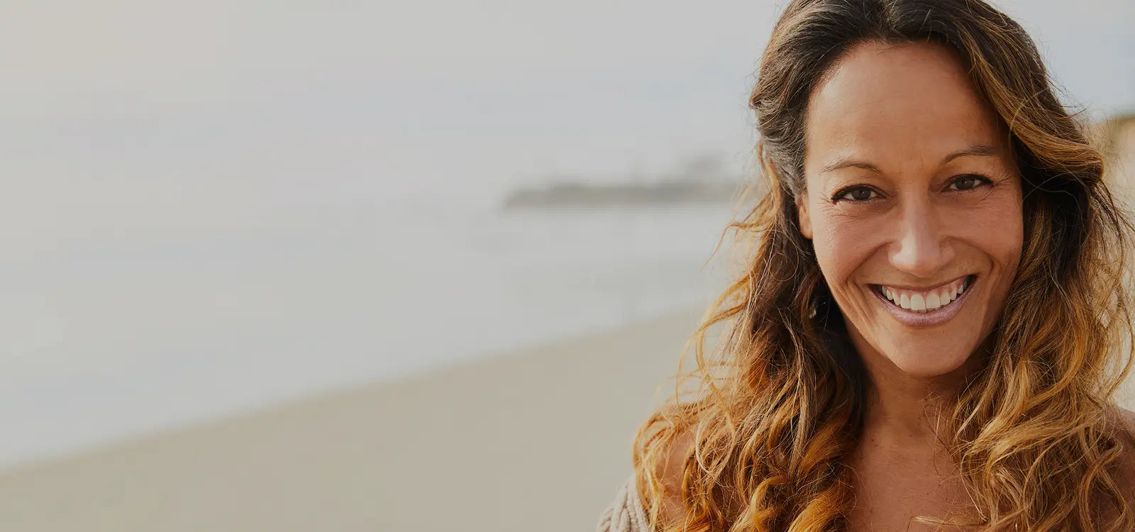 Close-up portrait of a happy, smiling middle-aged woman with beautiful, sun-kissed, wavy brown and blonde curly hair on a serene beach. She is looking directly into the camera, with a soft-focus background of sand and ocean.